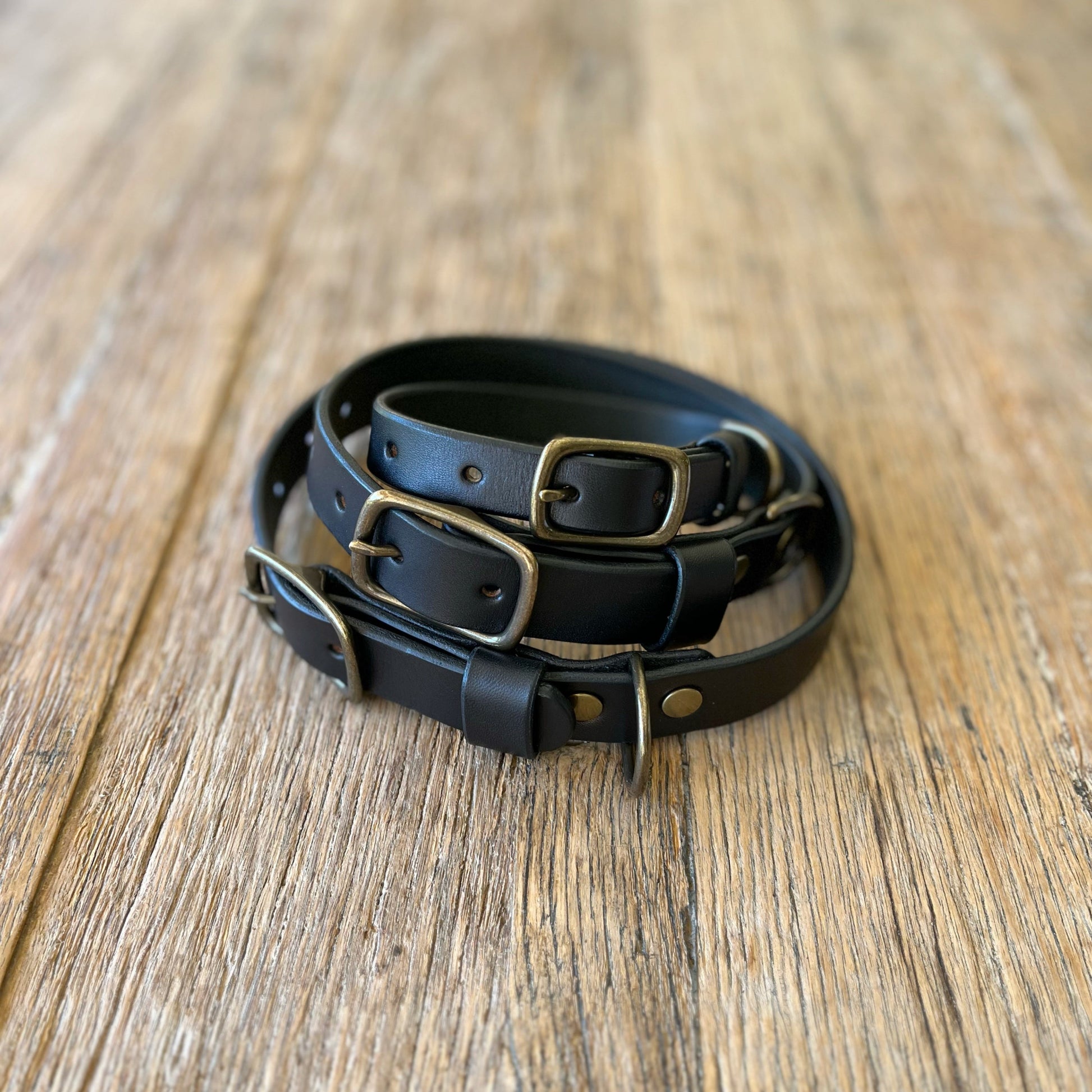 Three black leather dog collars with brass buckles on a wooden surface