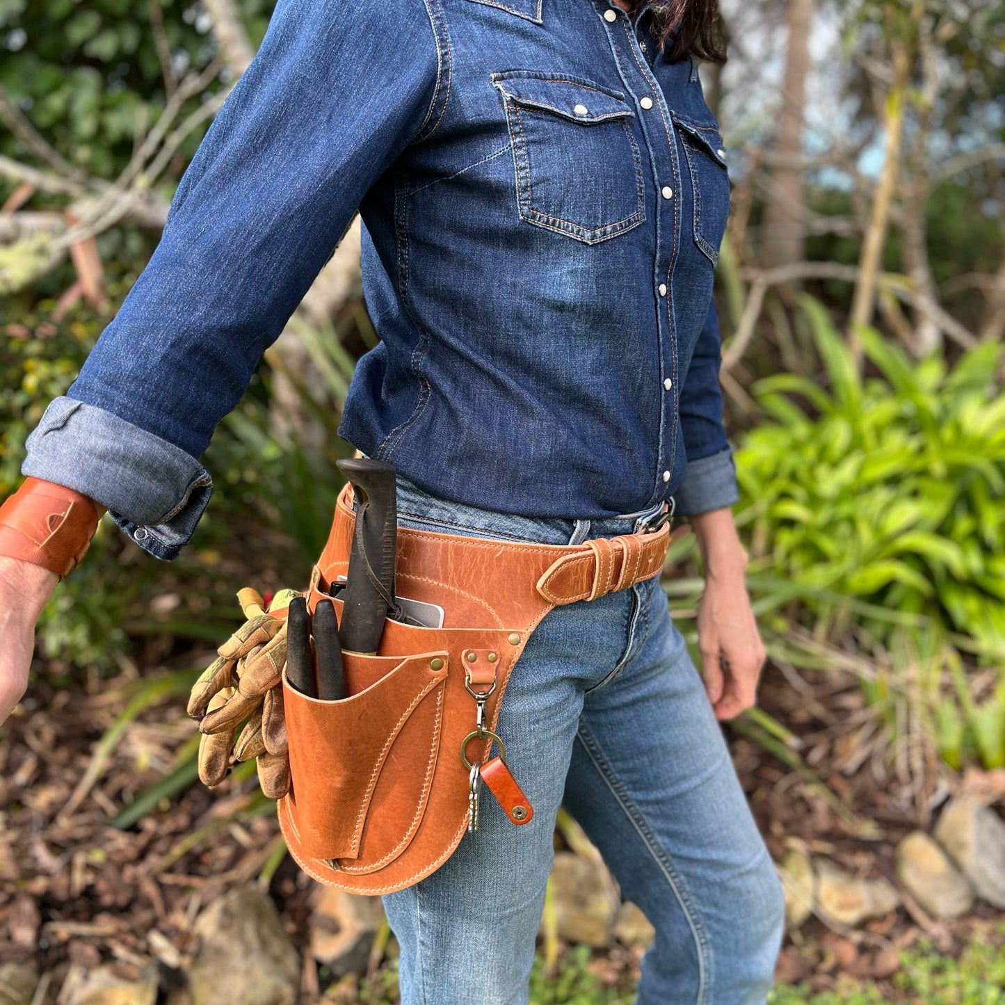 Person wearing a blue denim shirt and jeans with a brown leather tool belt outdoors.