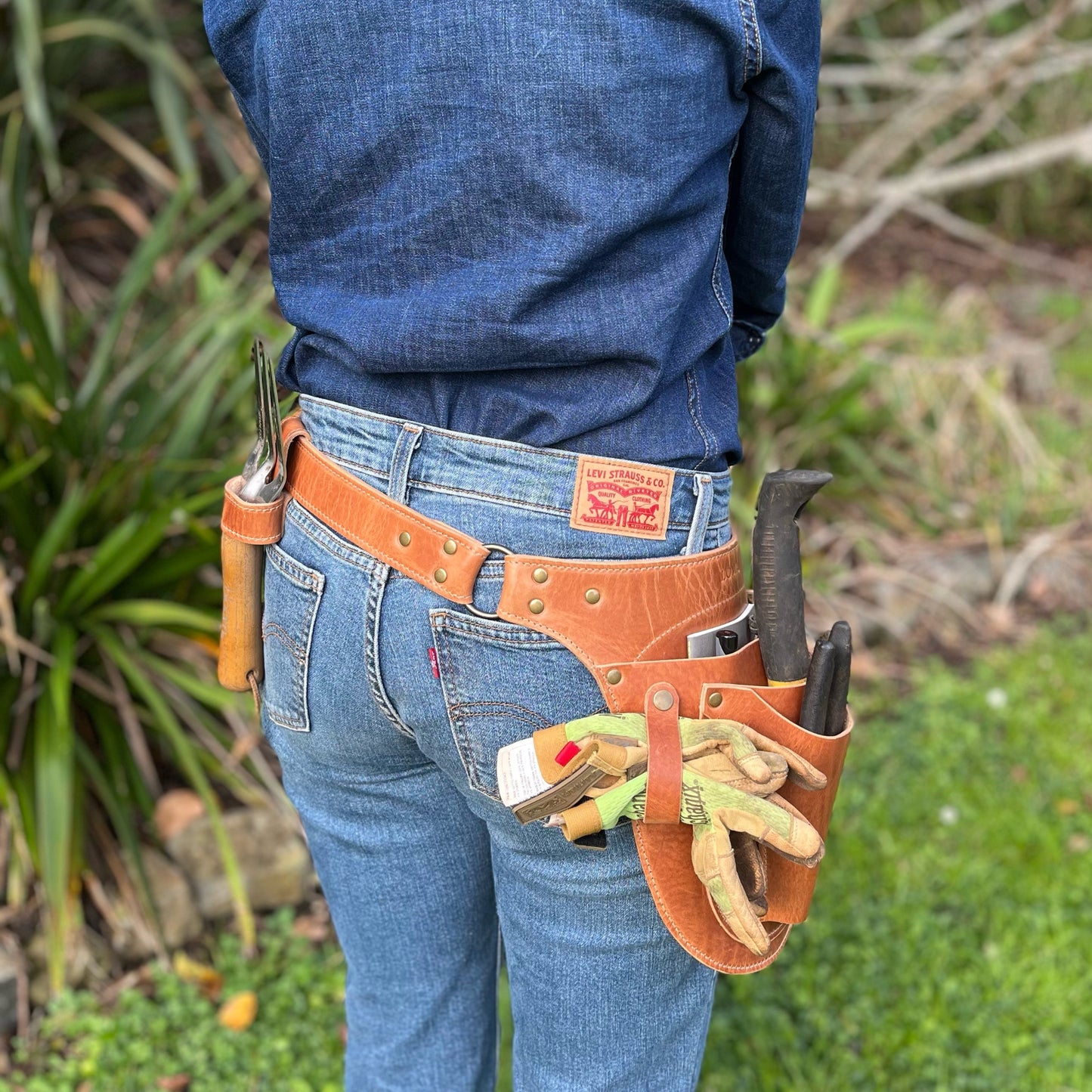 Person wearing a tool belt with various tools and gloves in an outdoor setting
