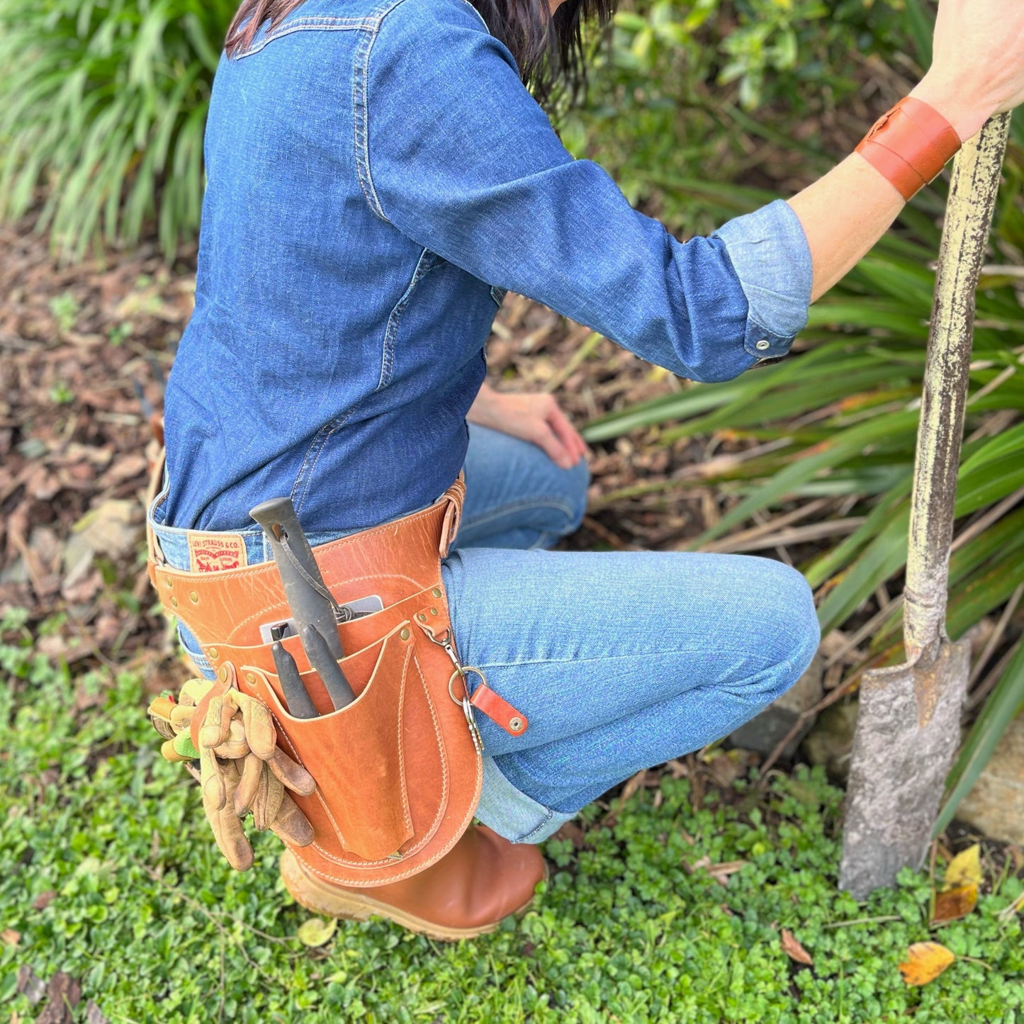 Person planting a tree with a tool belt on, surrounded by greenery