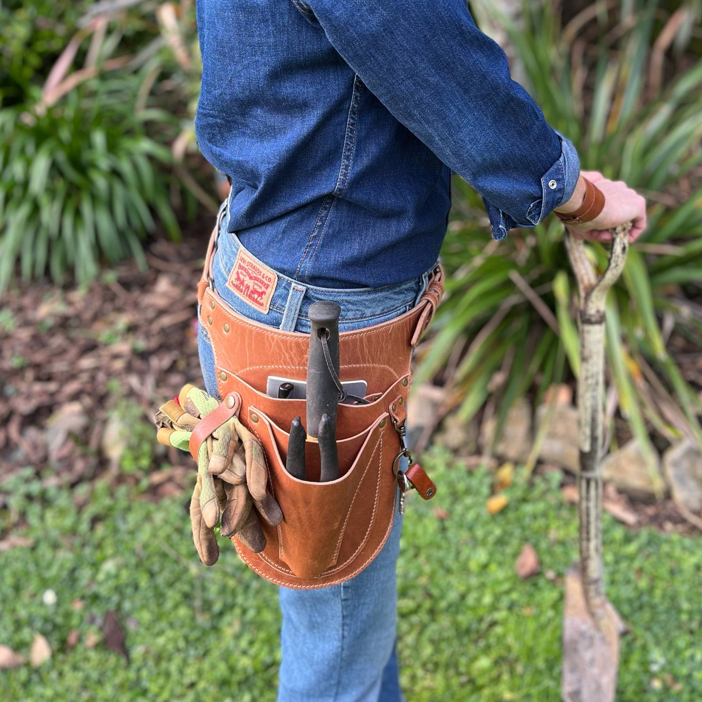 Person wearing a brown leather tool belt with gardening tools in an outdoor setting