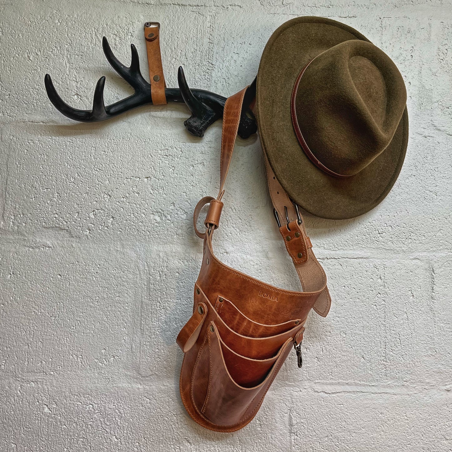 Brown leather tool belt and hat hanging on a wall with black antler hooks.