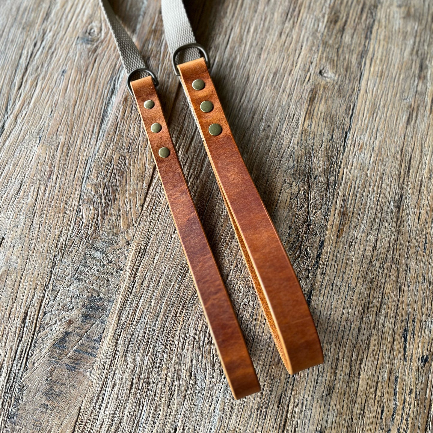 Two brown leather straps with brass rivets on a wooden surface