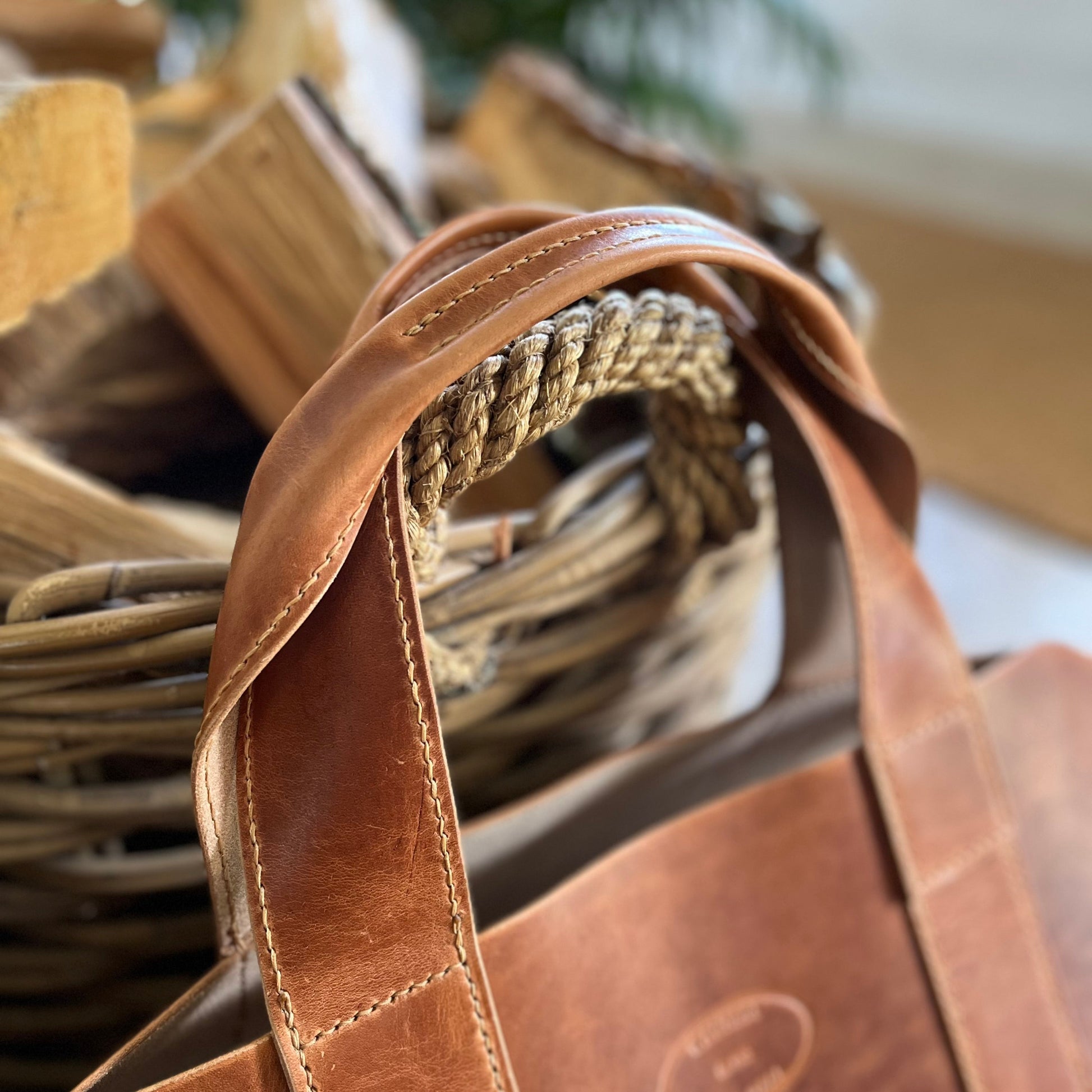 Close-up of a brown leather log carrier, resting in front of a wicker basket and stacked firewood in a warm indoor setting.