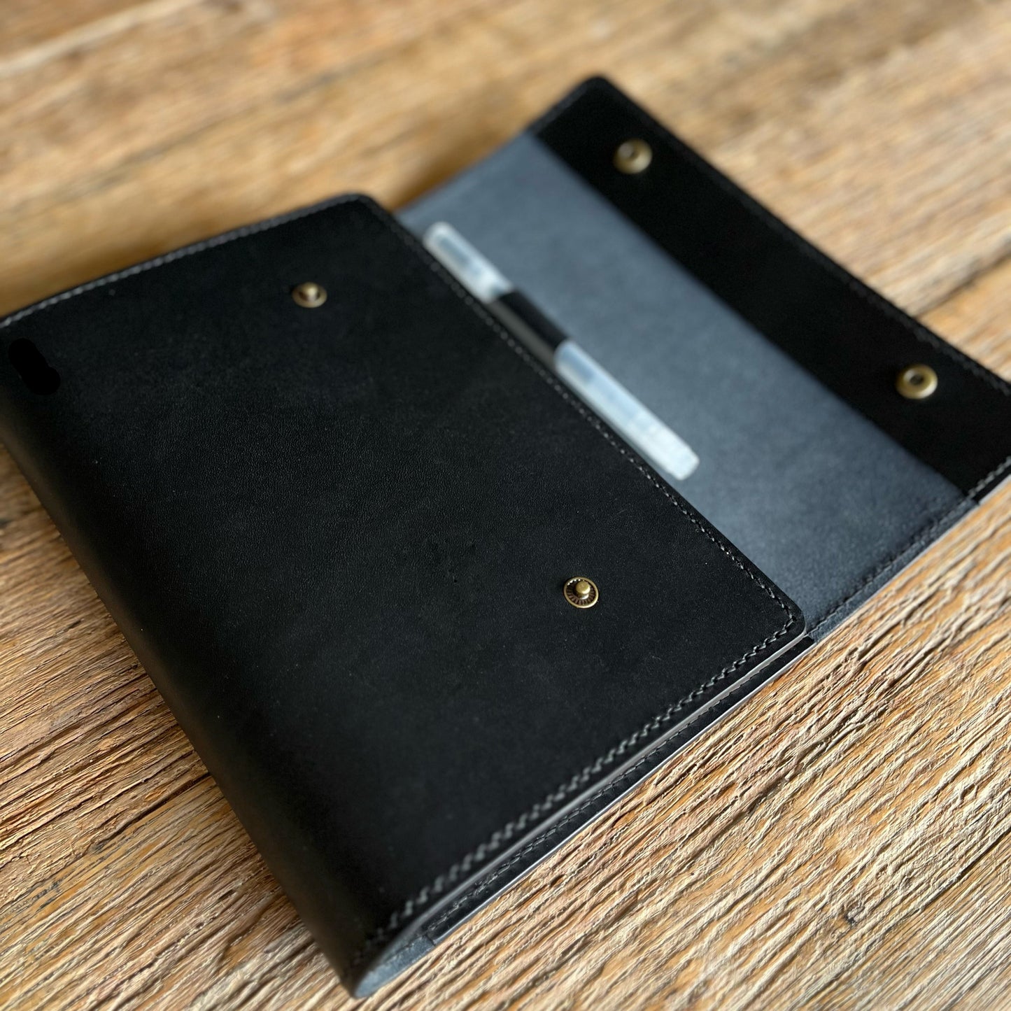 A black leather notebook cover or organizer, shown open on a rustic wooden table. The cover features brass snap buttons and a pen loop with a black and white pen inside.