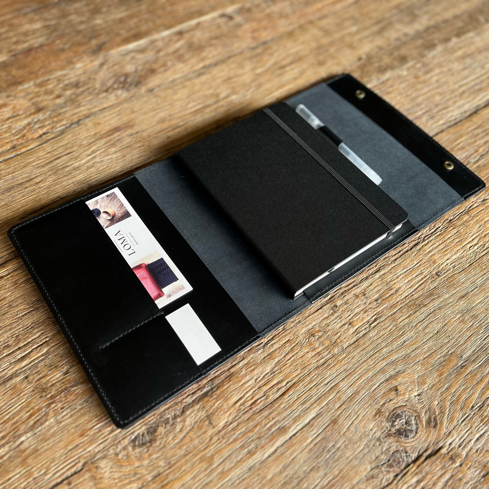 A black leather notebook cover and card holder organizer, shown open on a rustic wooden table. The organizer contains a black notebook, a pen in a slot, and sample cards.