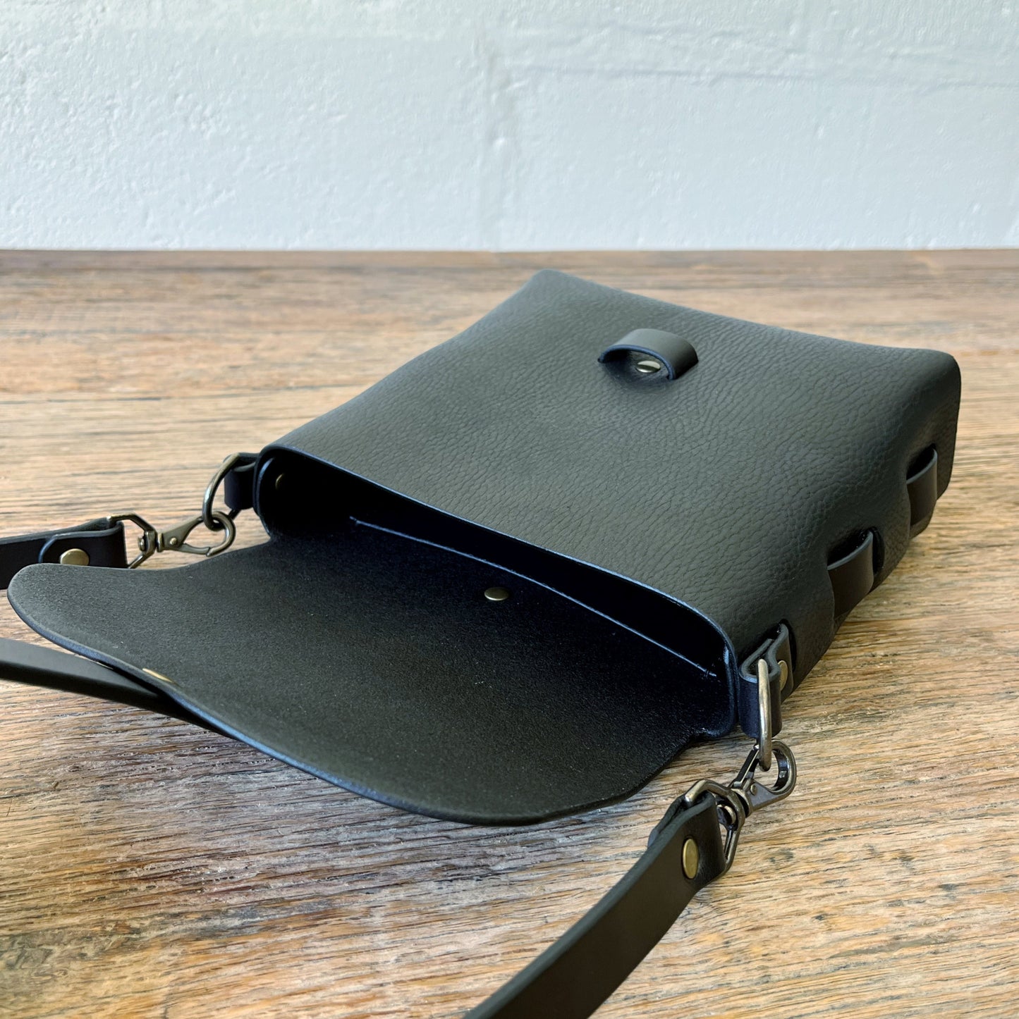 Black leather crossbody bag on a wooden surface with a white wall background