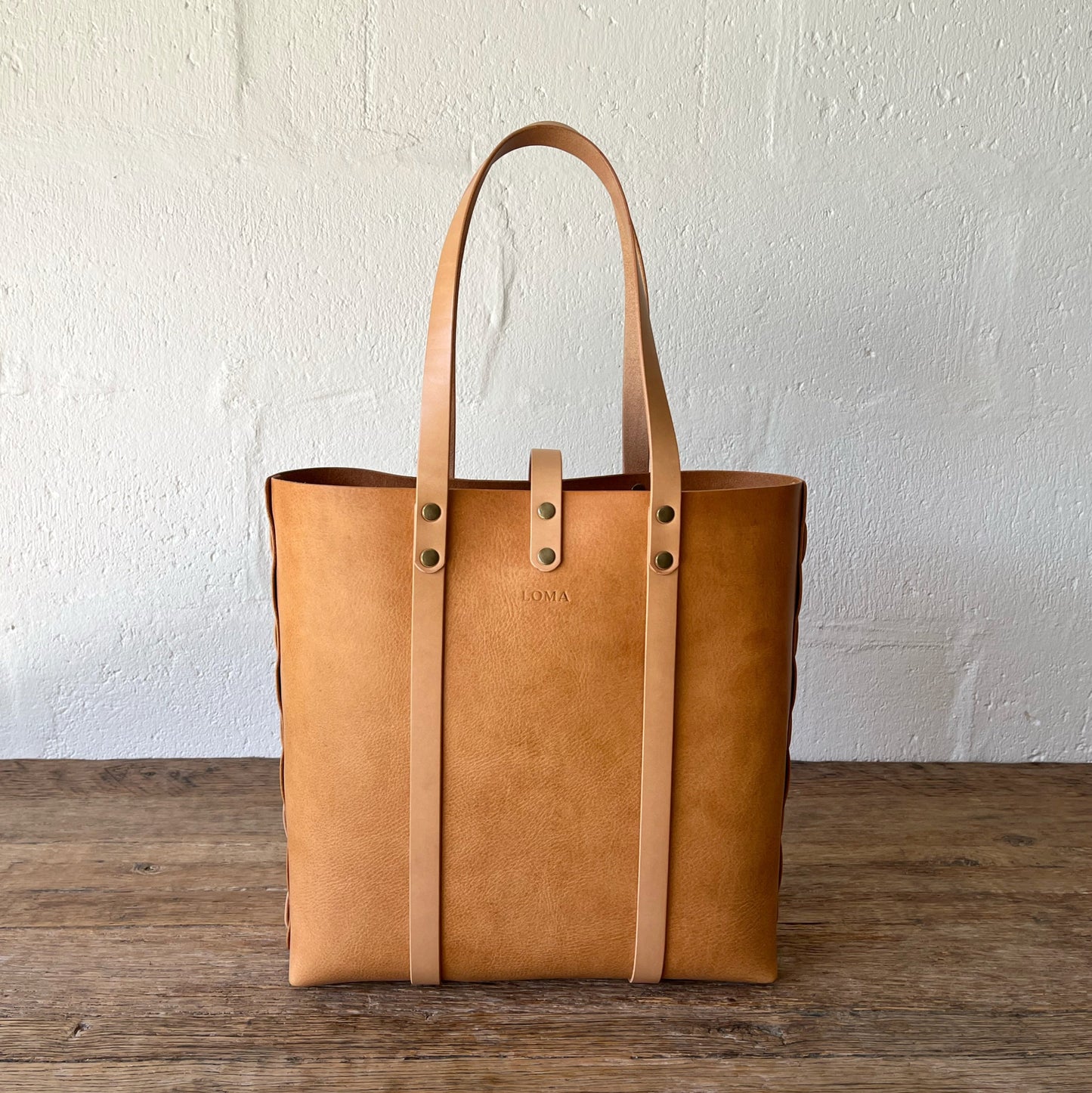 Brown leather tote bag on a wooden surface with a white textured wall background