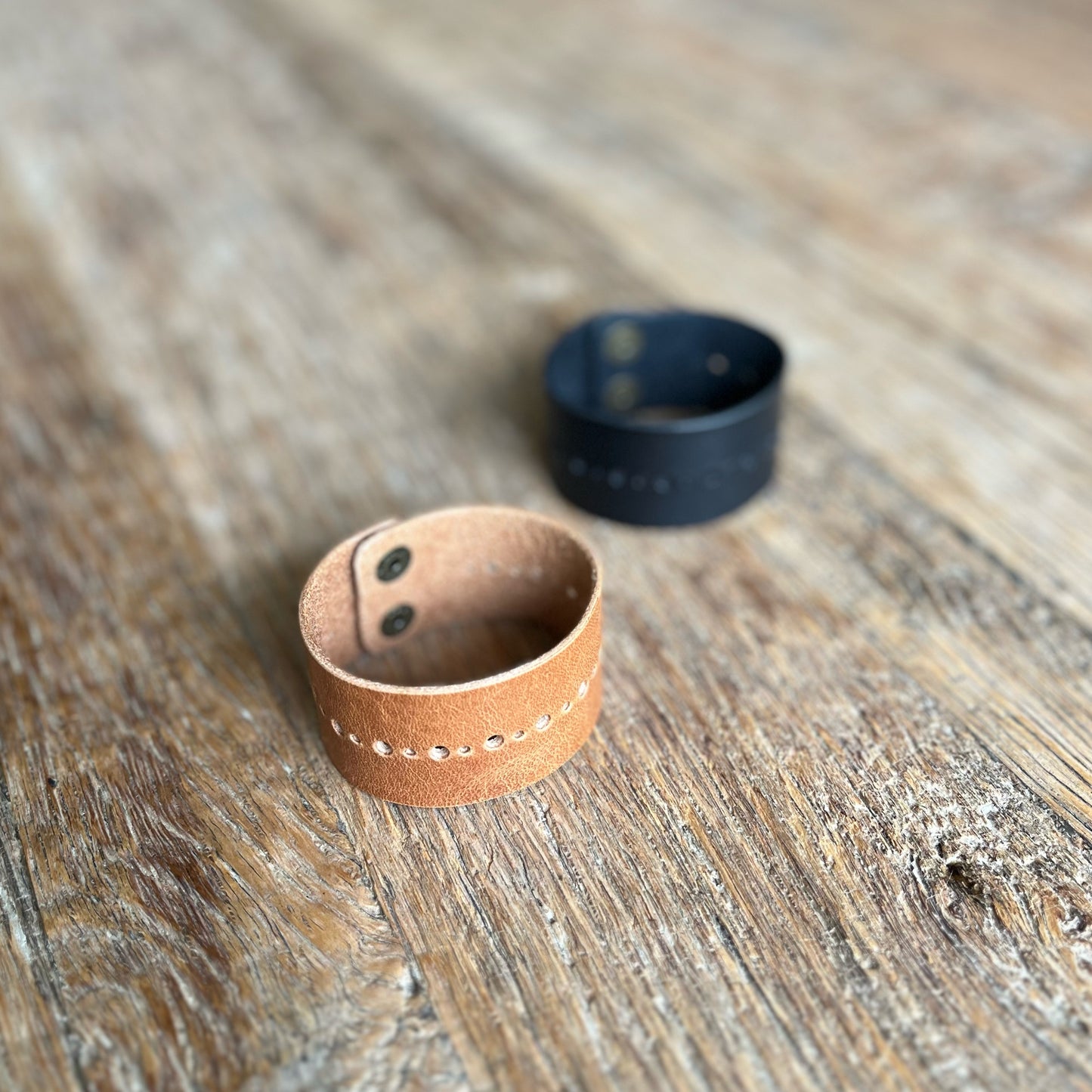 Two leather cuffs on a wooden surface, one in black and one in brown, with perforated dot details and brass snap buttons.
