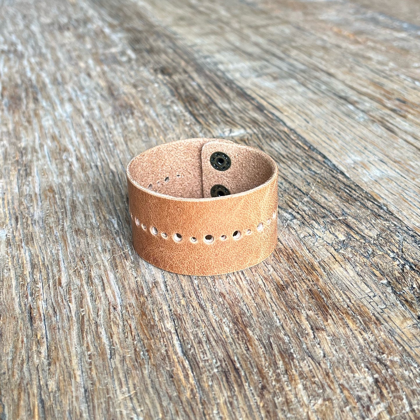 leather cuff on a wooden surface, with perforated dot details and brass snap buttons.