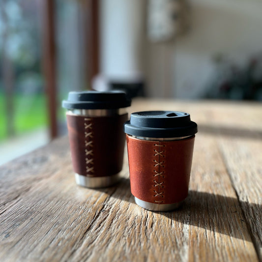 Two brown leather coffee cups with black lids on a wooden surface.