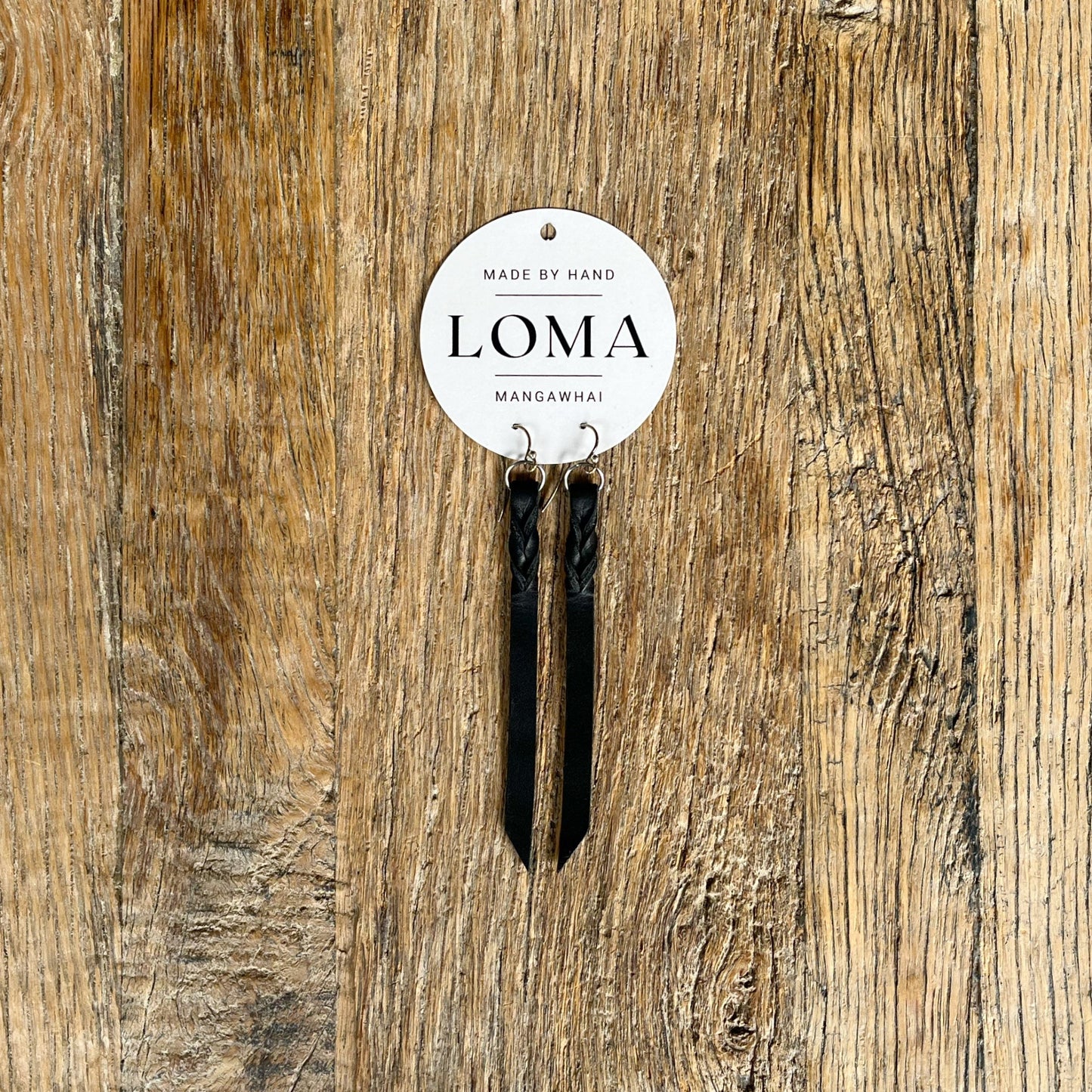Black leather twist earrings on a wooden surface with a 'LOMA' label.