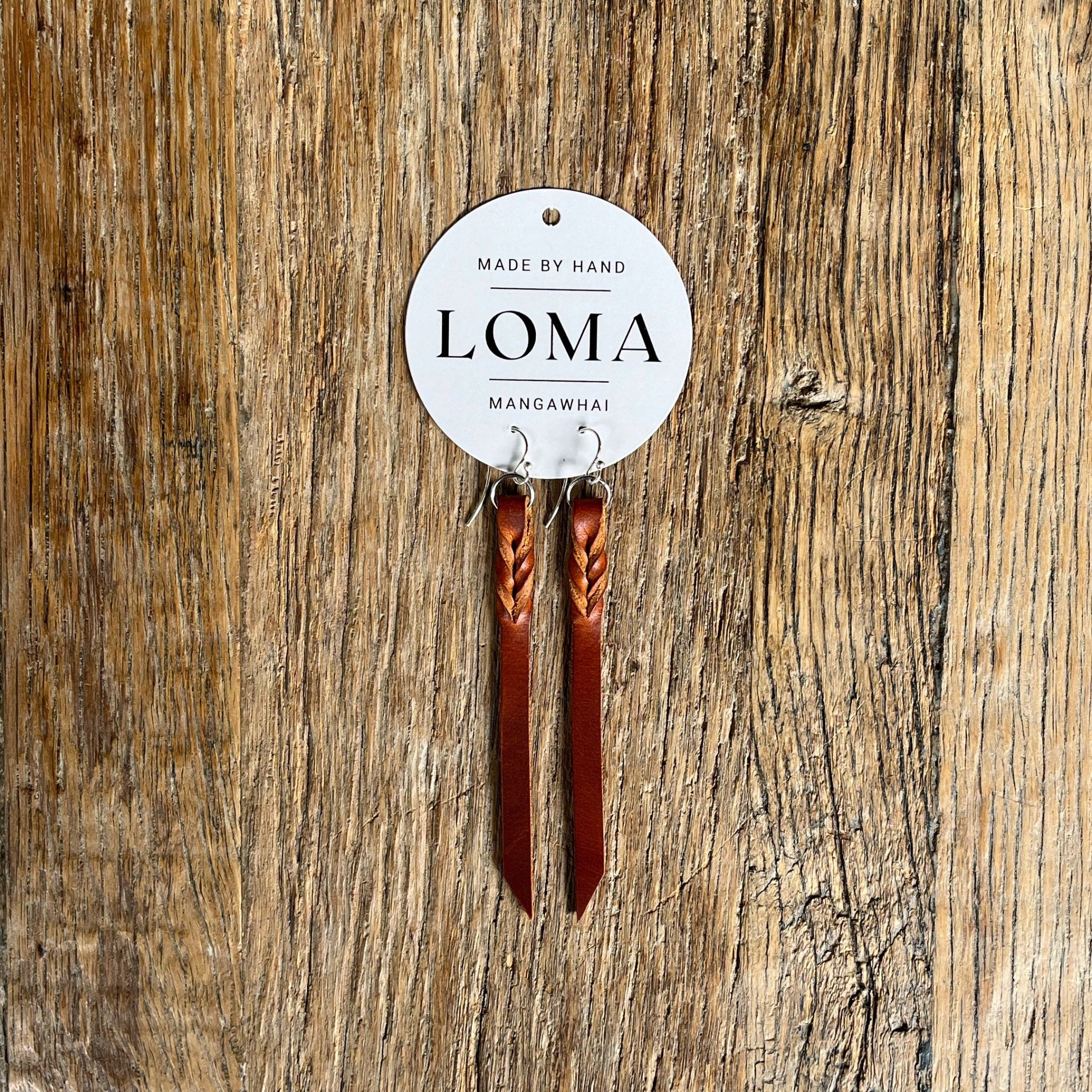 Brown leather earrings with twist design on a wooden surface, featuring a 'LOMA' label.