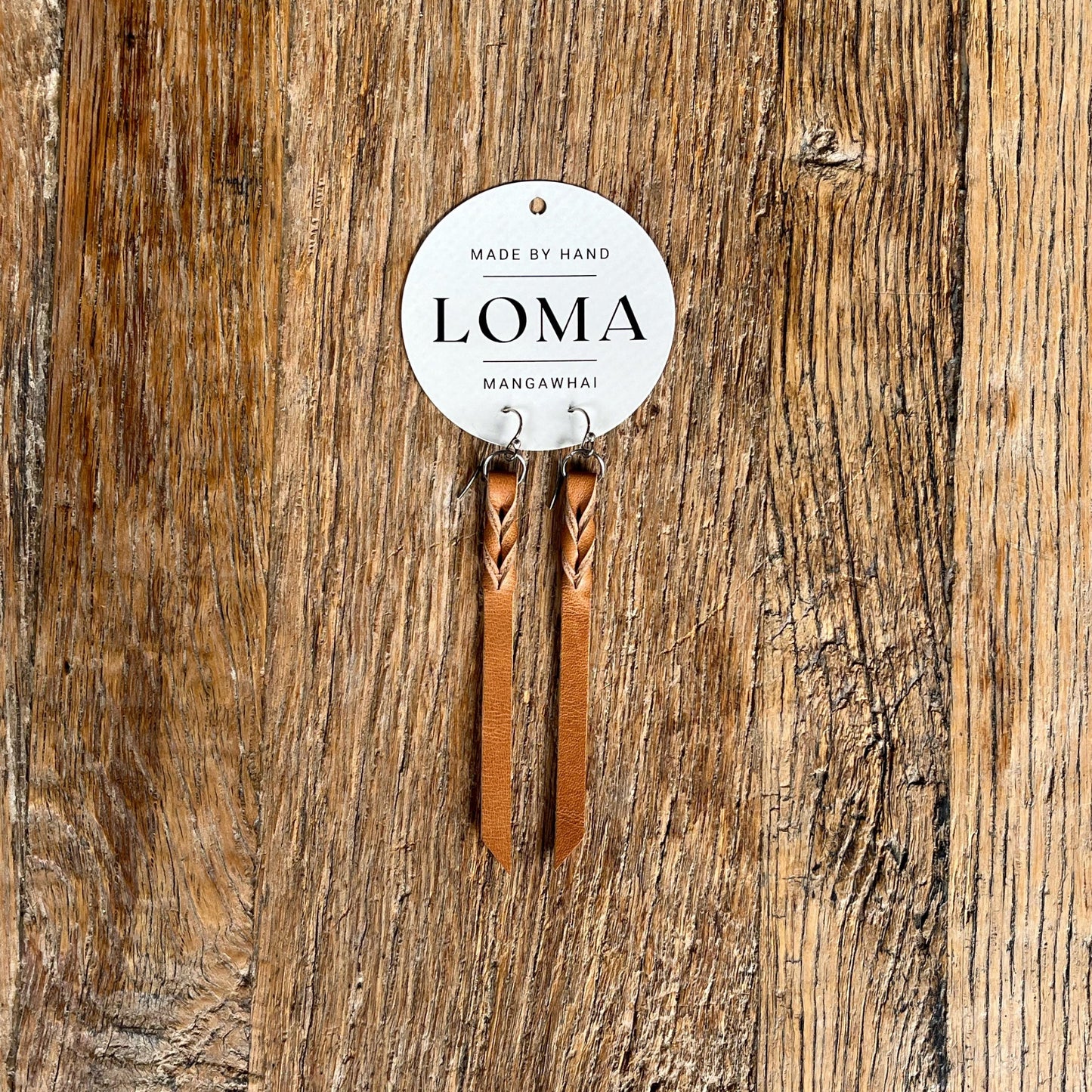 Pair of natural coloured leather earrings with a 'LOMA' label on a wooden background