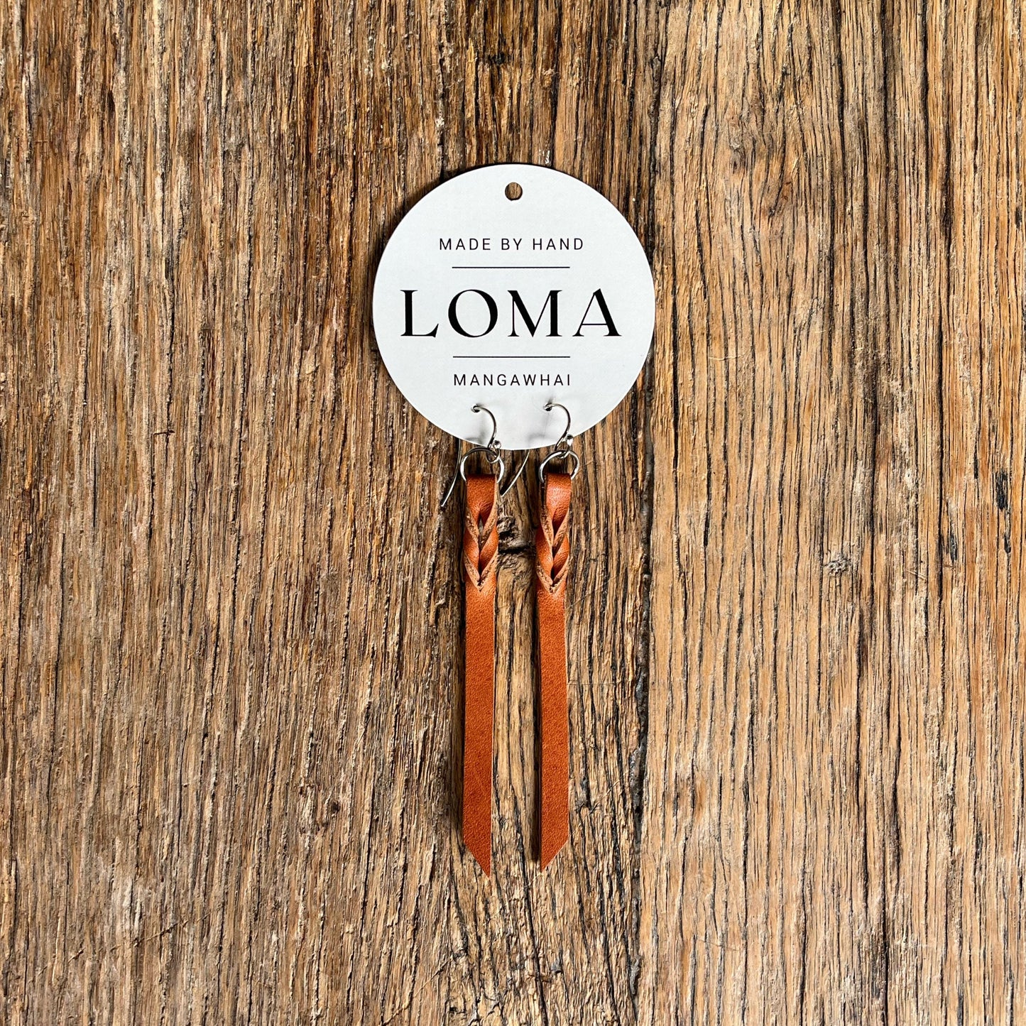 Tan leather earrings on a 'LOMA' branded card against a wooden background