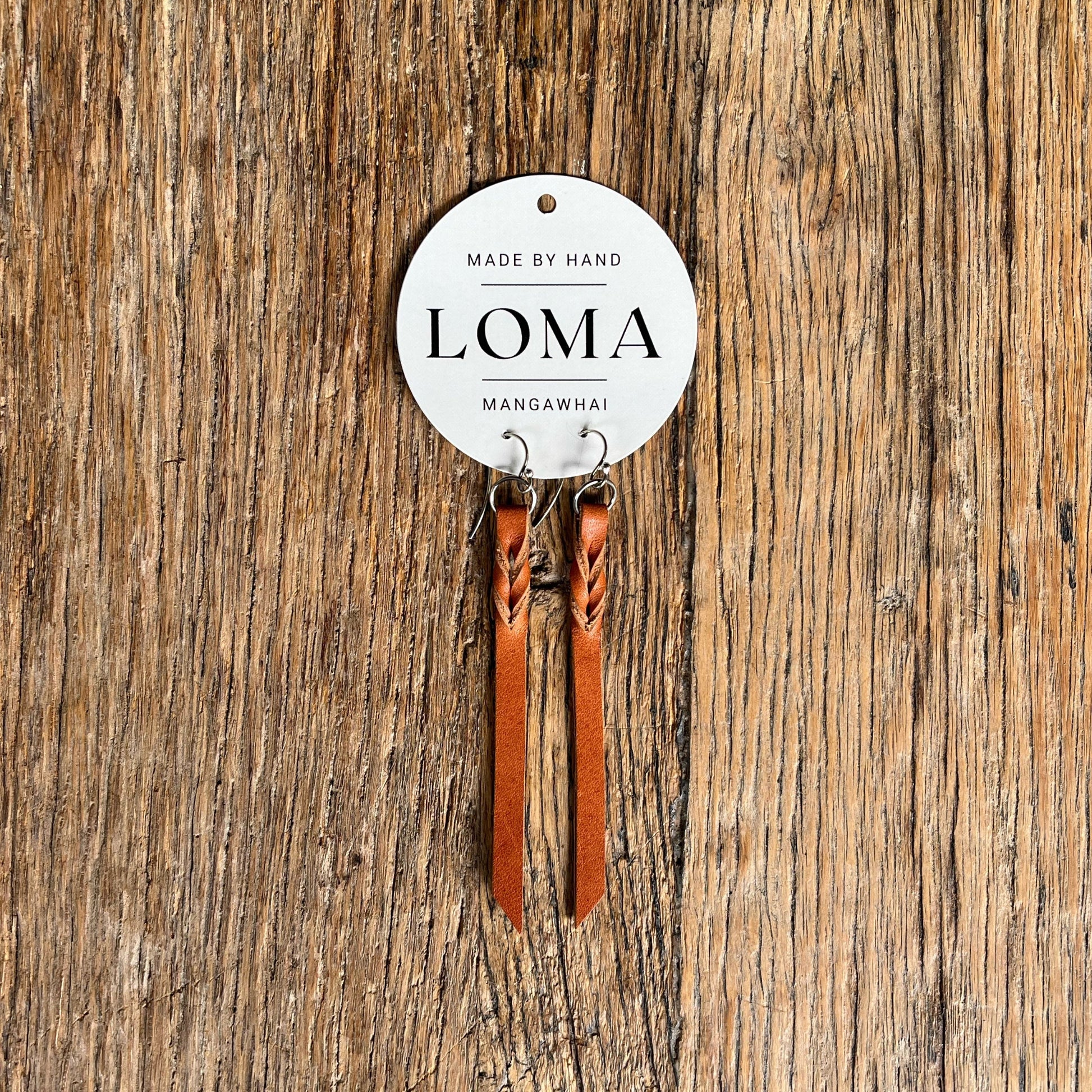 Tan leather earrings on a 'LOMA' branded card against a wooden background