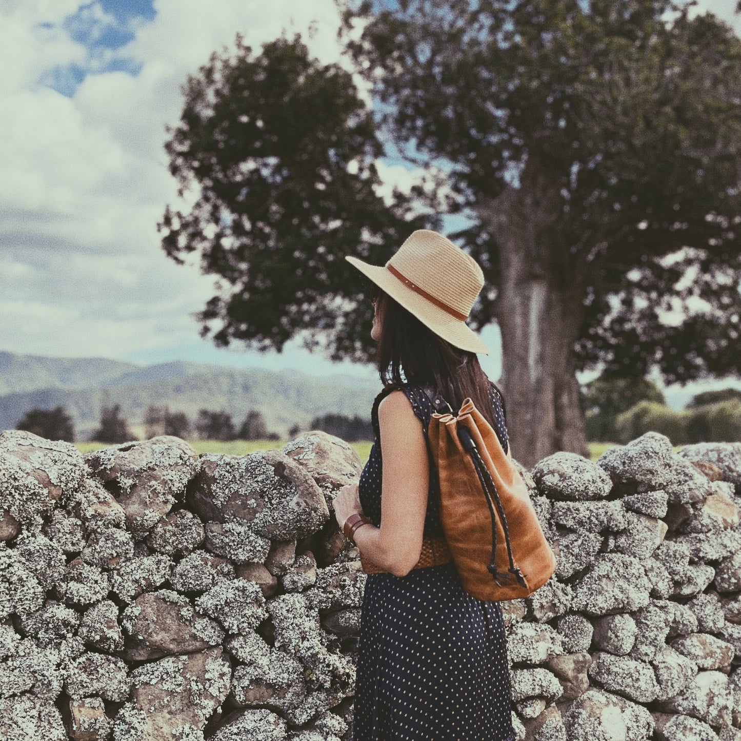 Woman with a straw hat and brown backpack standing near a stone wall with trees and mountains in the background.