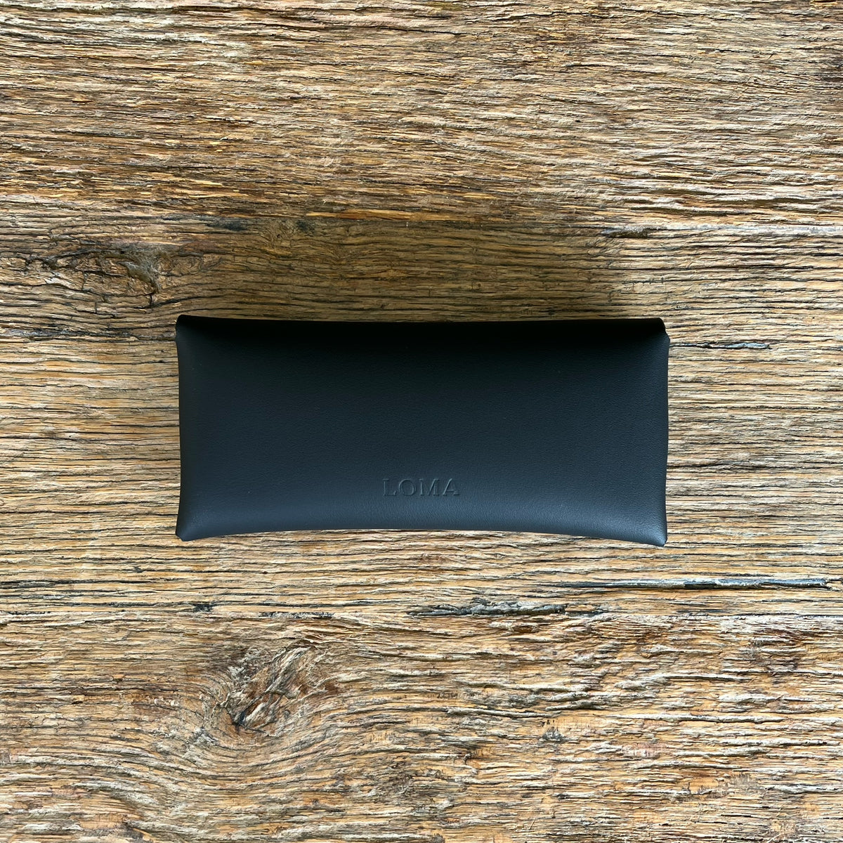 A black leather eyeglasses case with debossed 'LOMA' branding, placed on a rustic, textured wooden surface.
