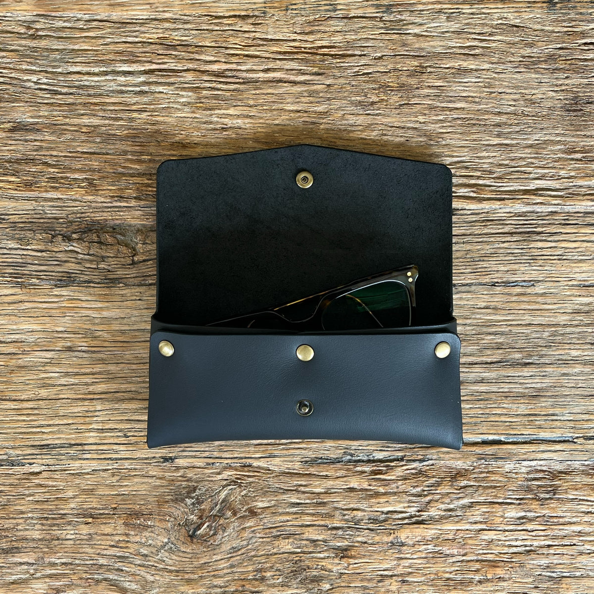 A black leather sunglasses case with brass snap buttons, shown open on a rustic wooden surface with sunglasses inside.