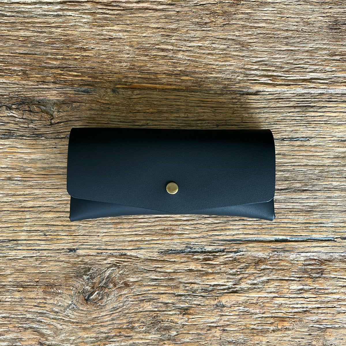 A black leather glasses case with a small brass snap button, laid flat on a rustic wooden surface.