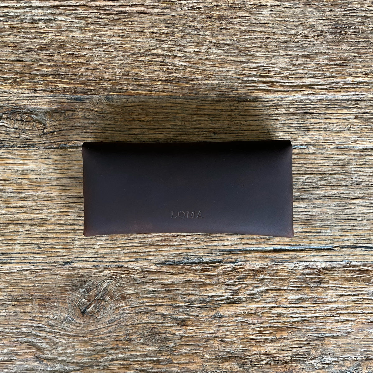 A dark brown leather glasses case with the brand name 'LOMA' embossed on the front, resting on a rustic wooden surface.