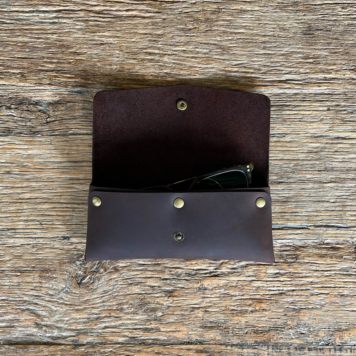 A dark brown leather sunglasses case with brass-tone rivets, shown open on a rustic wooden surface. A pair of sunglasses is partially tucked inside.