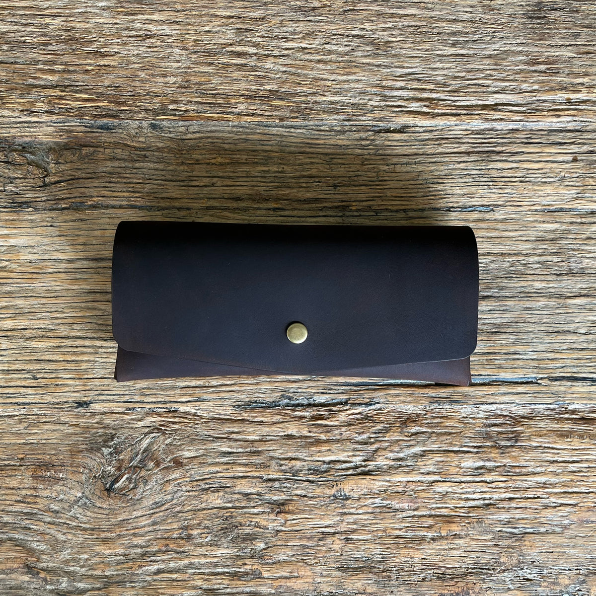 A dark brown leather glasses case with a small gold snap button, placed on a rustic wooden surface.