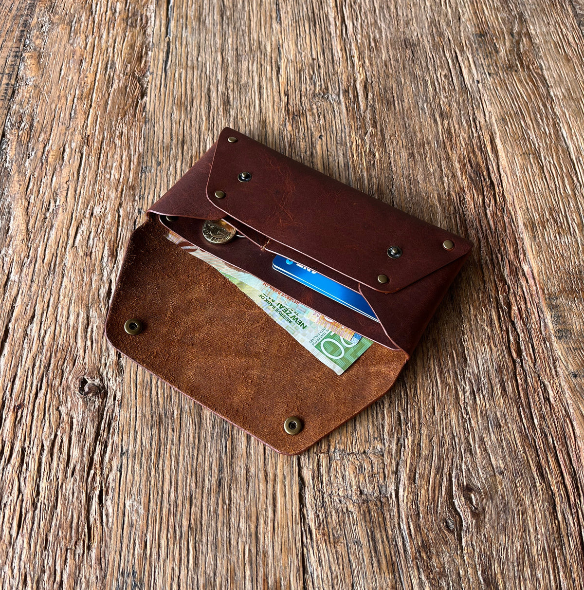 A dark brown leather wallet with brass snaps, shown open on a rustic wooden surface with cash and cards visible inside.