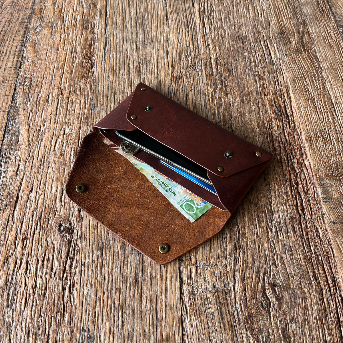 A brown leather card holder wallet with an envelope-style flap, shown open on a rustic wooden surface. Inside, a smartphone and cards are partially visible.