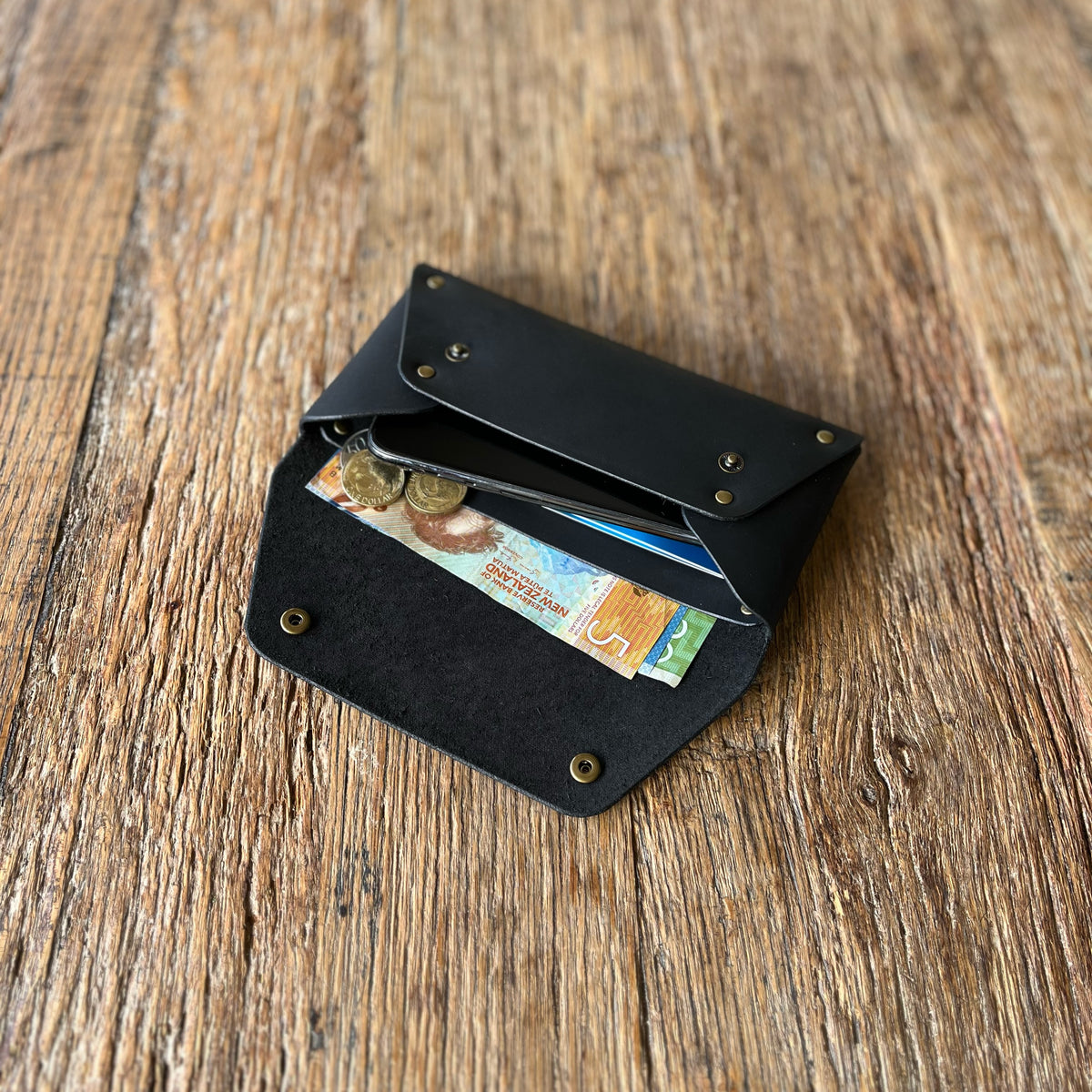 A black leather wallet with brass snap buttons, shown open on a rustic wooden surface, containing cards, cash, and a coin.