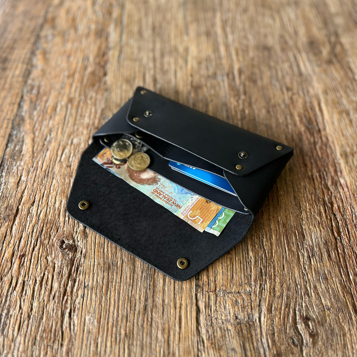 A black leather wallet with brass snaps, shown open on a rustic wooden surface, containing cash, coins, and a card.