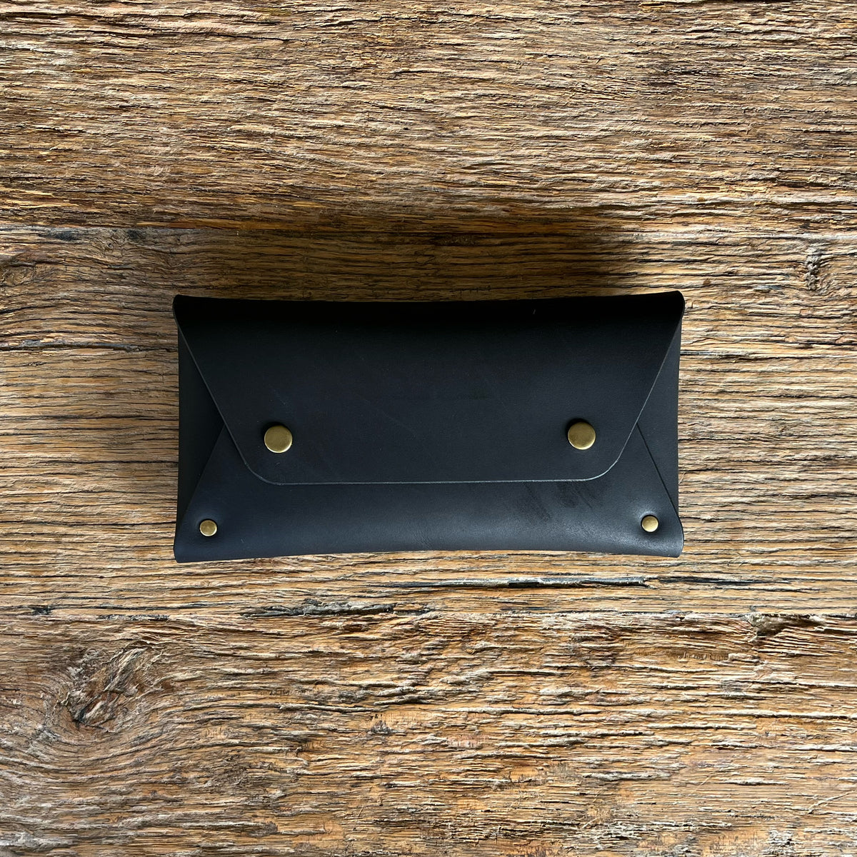 A black leather purse with a flap closure and brass snap buttons, displayed flat on a rustic wooden surface.