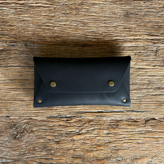 A black leather purse with a flap closure and brass snap buttons, displayed flat on a rustic wooden surface.