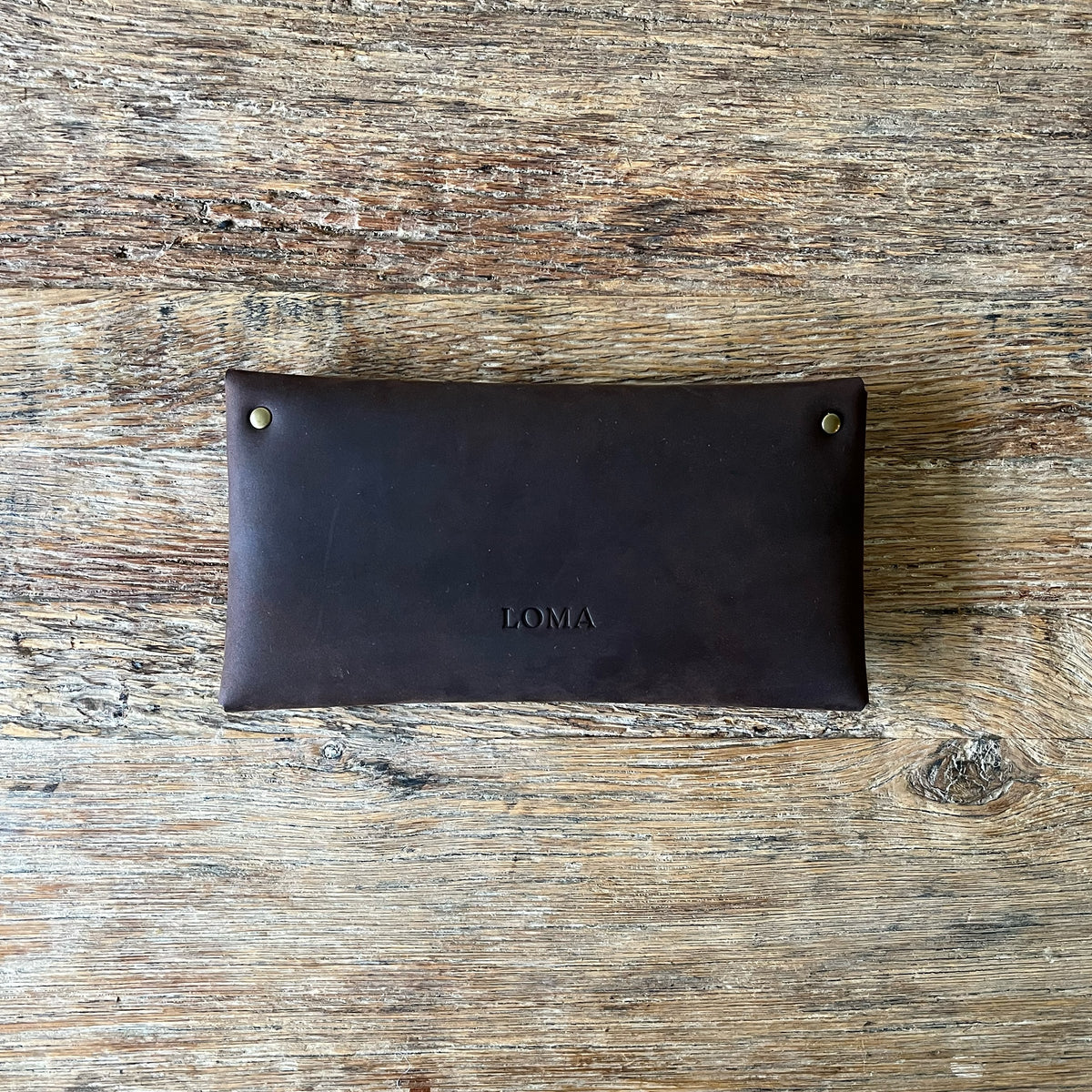 A dark brown leather pouch with brass grommets and the brand name 'LOMA' embossed on the front, laid flat on a rustic wooden surface.