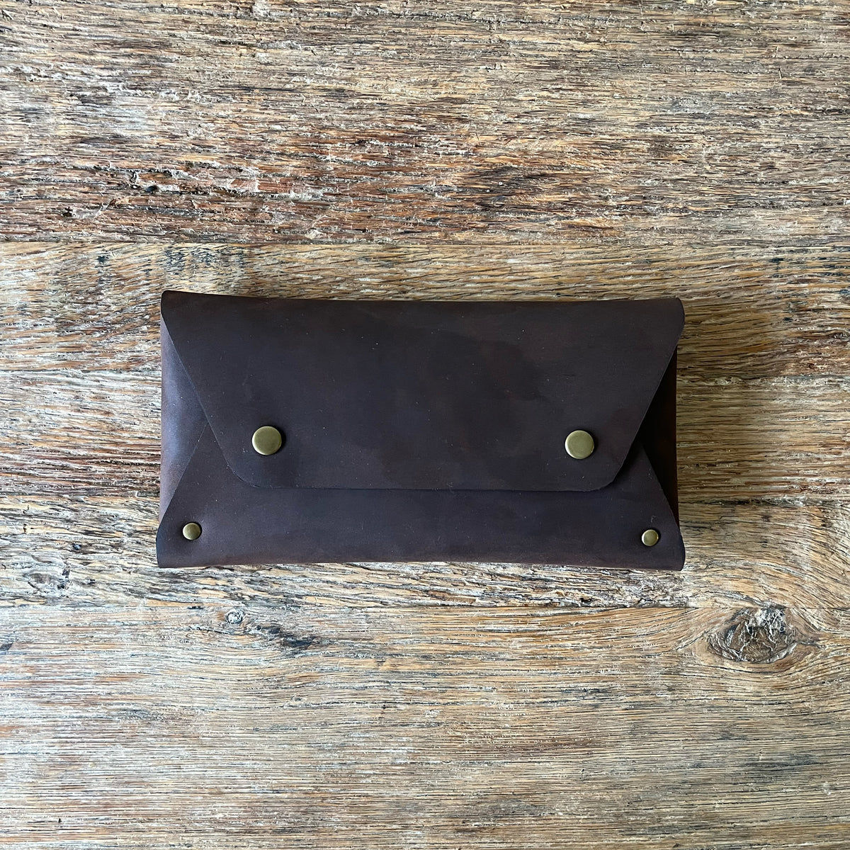 A dark brown leather pouch with brass snap buttons, laid flat on a rustic wooden surface.