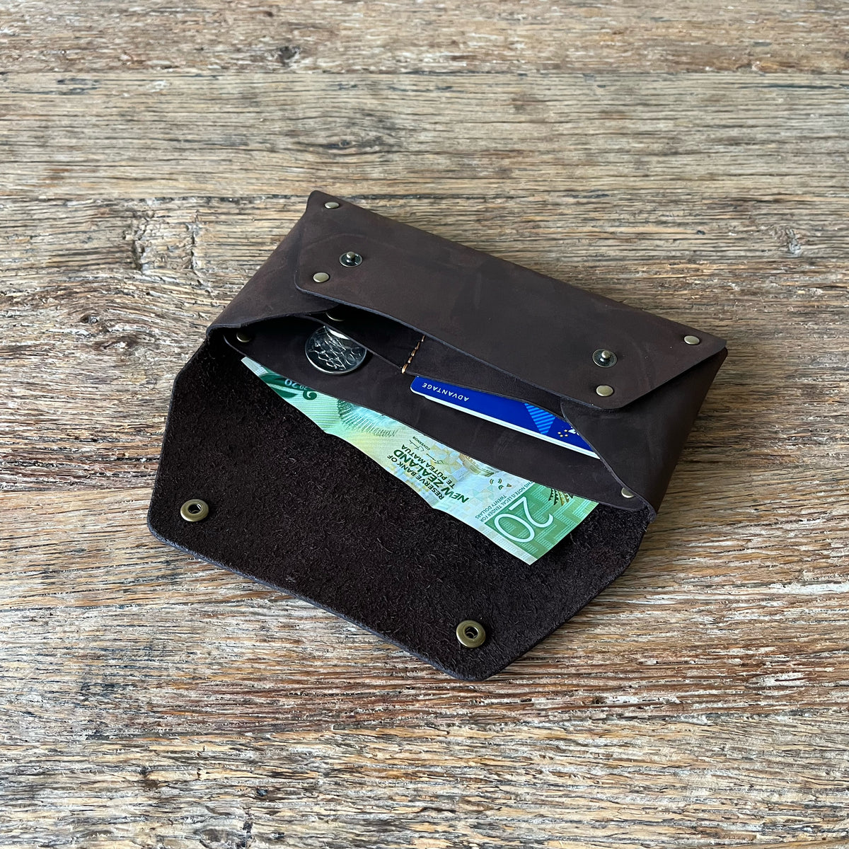 A dark brown leather card holder wallet with a flap closure, shown open on a rustic wooden surface. The wallet contains cash, a coin, and a blue credit card.