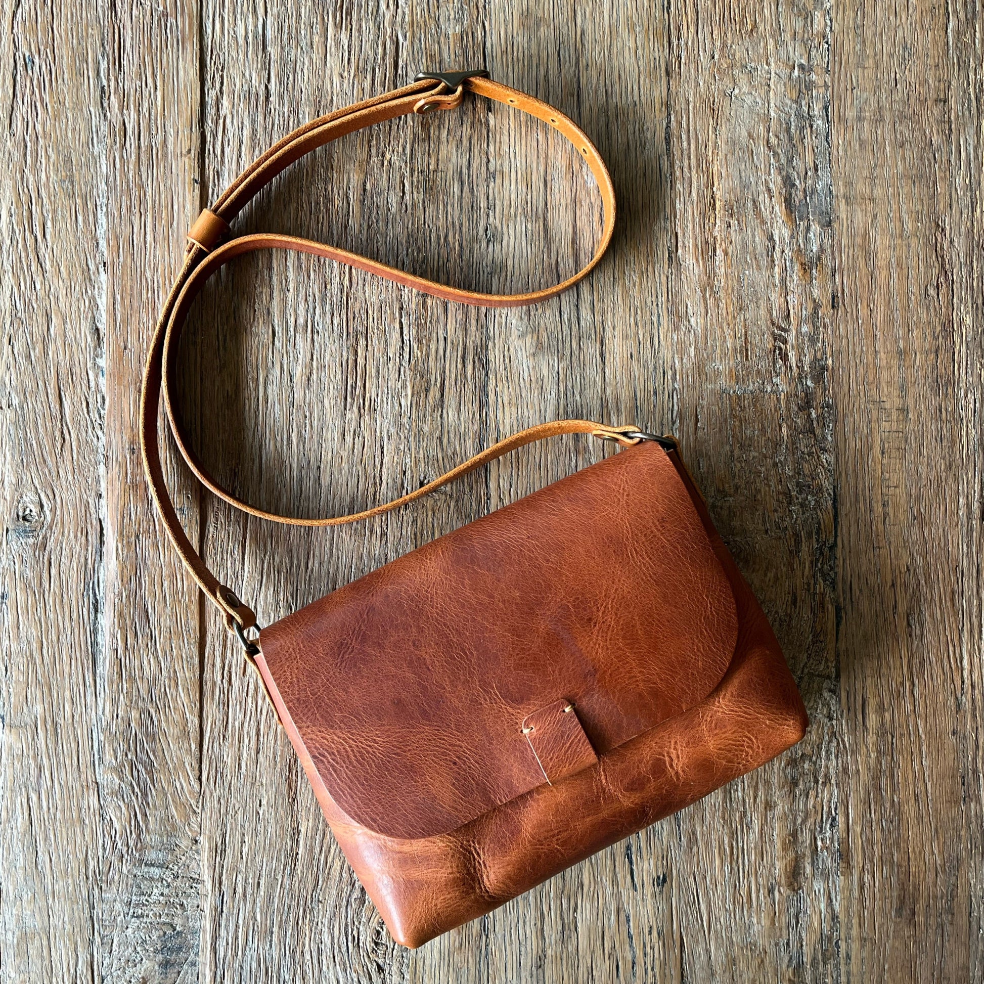 A brown leather shoulder bag with a long adjustable strap, laid flat on a rustic wooden surface.