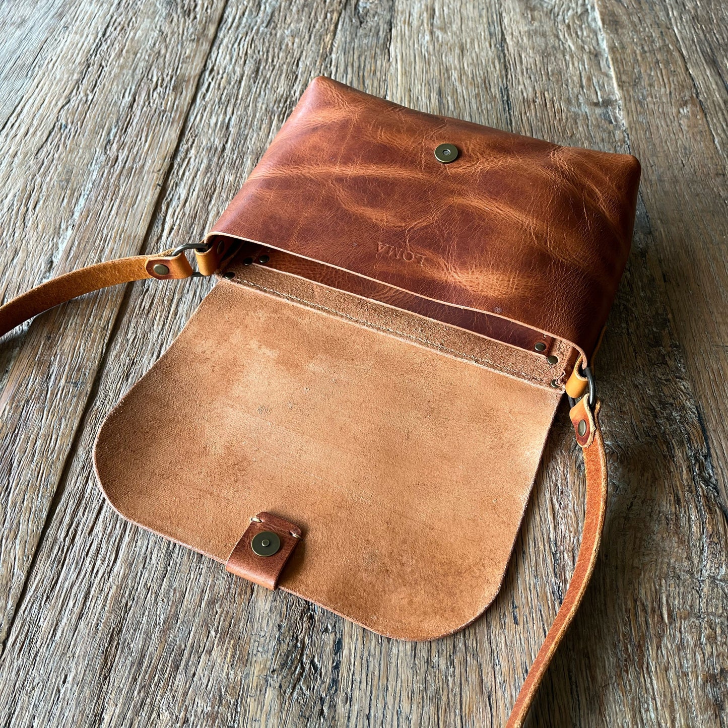 A brown leather crossbody bag with a flap closure, shown open on a rustic wooden surface. The bag features a darker brown textured leather exterior and a lighter tan leather interior, with a matching leather strap and small brass-tone snap button.