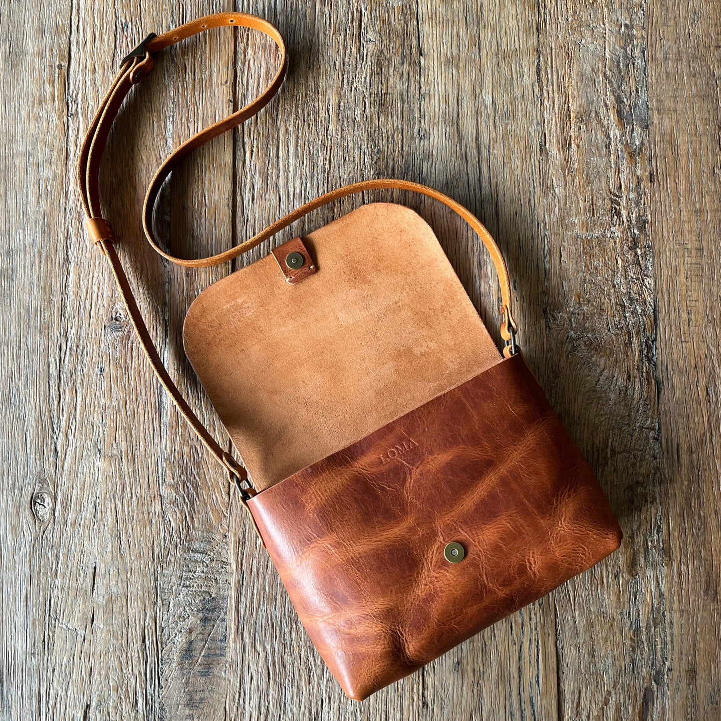 A brown leather crossbody bag with a long strap, laid flat on a rustic weathered wood background.