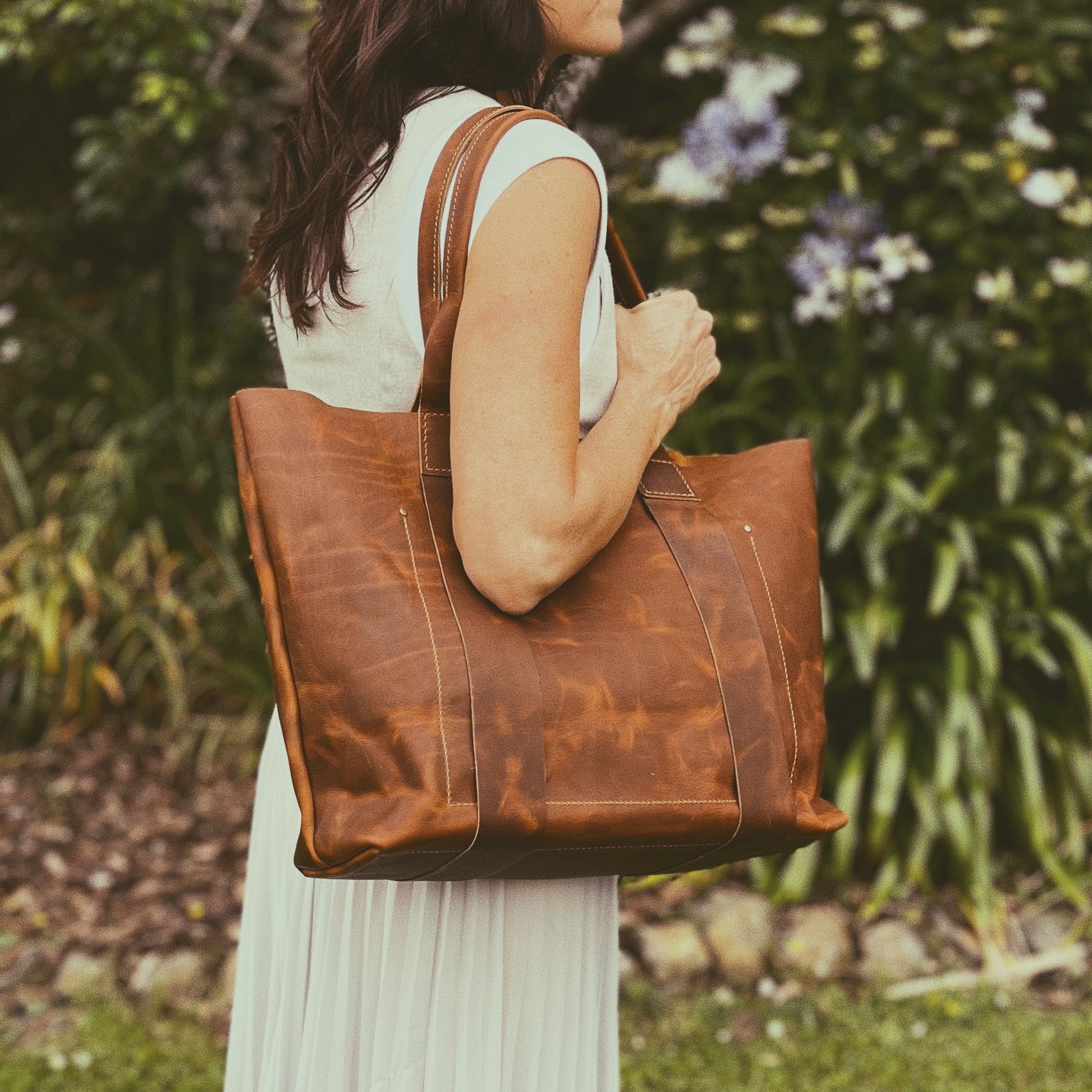 Person holding a brown leather tote bag outdoors with greenery in the background