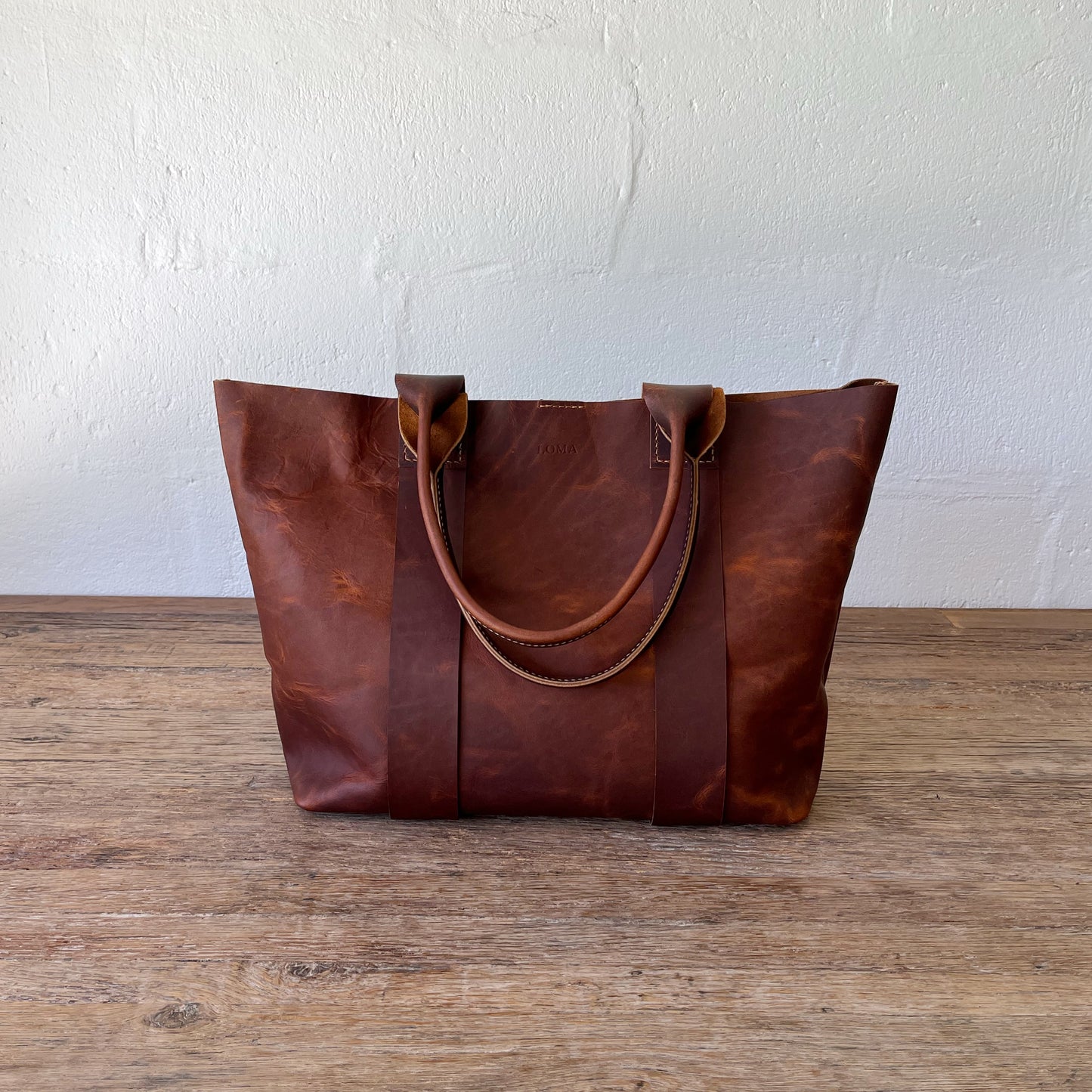 Brown leather tote bag on a wooden surface with a white wall background