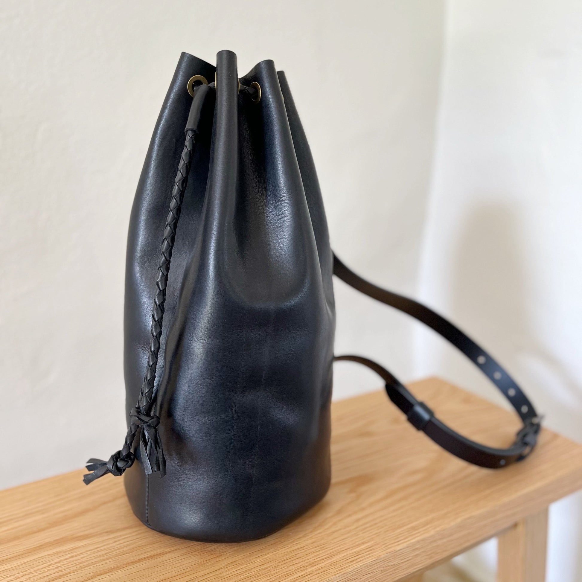 Black leather bucket bag with a strap on a wooden surface against a white background