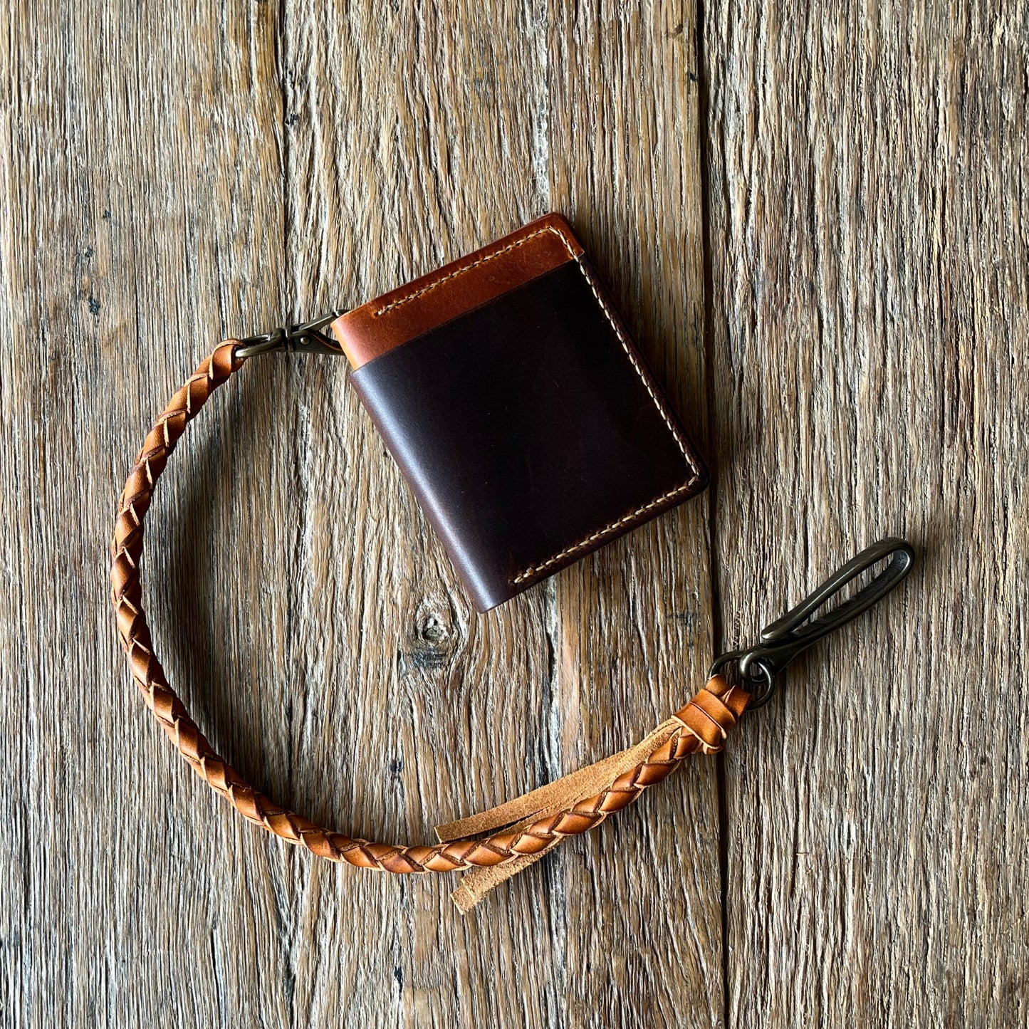 A brown bifold cash wallet with a hand braided leather lanyard and brass lever clip, placed on a wooden surface.