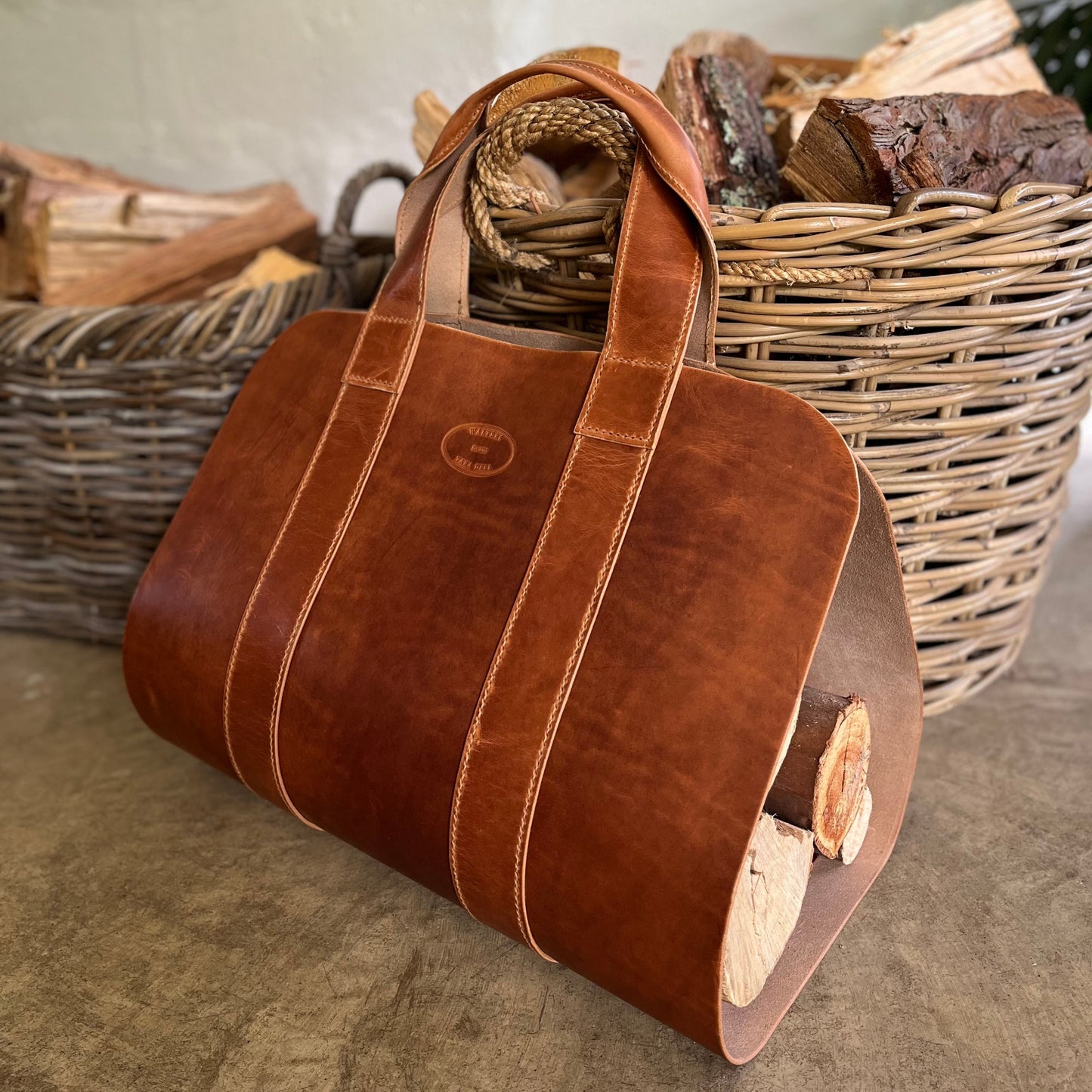 A brown leather log carrier with double top handles, shown in a rustic setting with wicker baskets of firewood in the background.